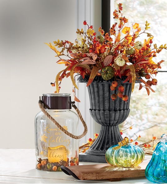 Fall tabletop scene in front of a window features a floral urn topiary with autumn leaves and colors and 3 colored glass pumpkins.